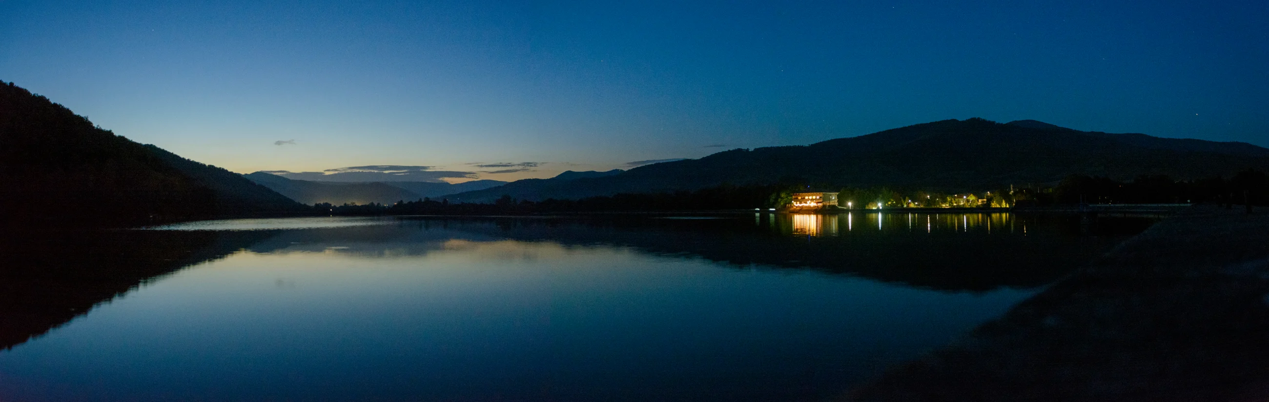 Wide panorama of a lake, hills and reflected lights at blue hour